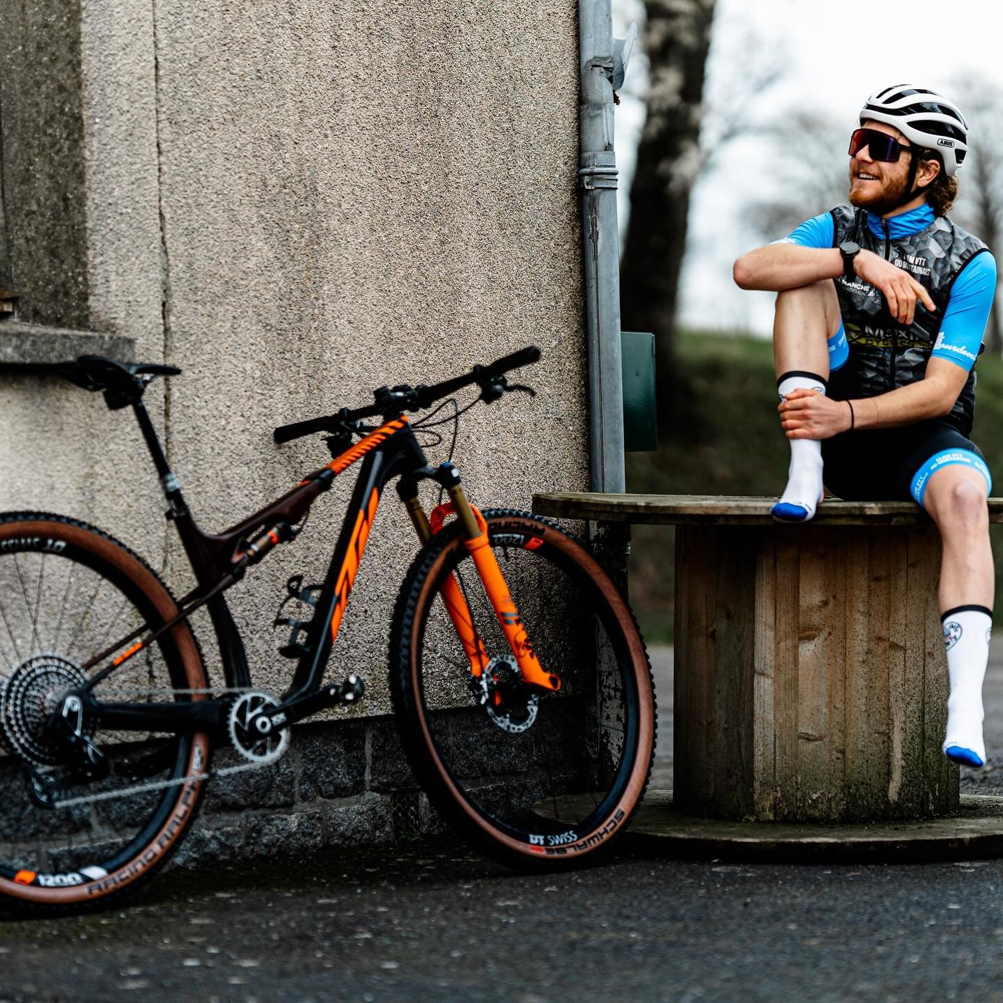 Cycliste en tenue de VTT assis sur un banc, portant des chaussettes Cycling and Sports, à côté d’un VTT KTM tout-terrain orange appuyé contre un mur.