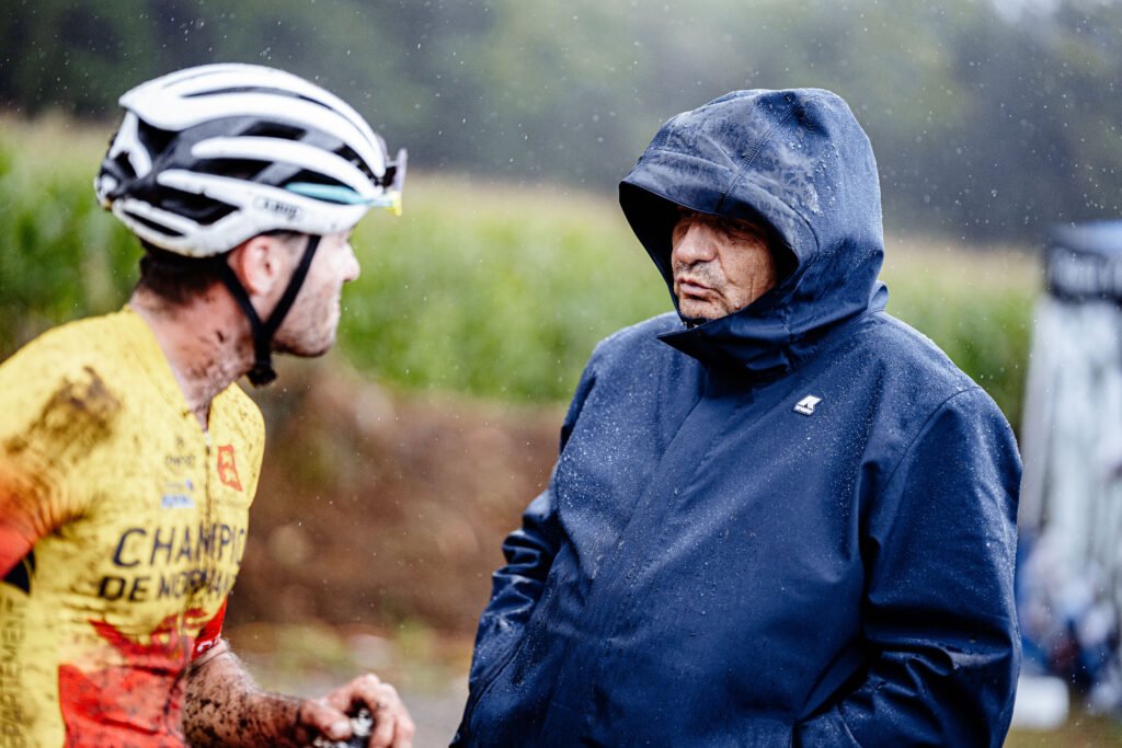 Laurent Hirtz échange avec un cycliste couvert de boue sous la pluie, lors d’une course ou d’un entraînement en extérieur.