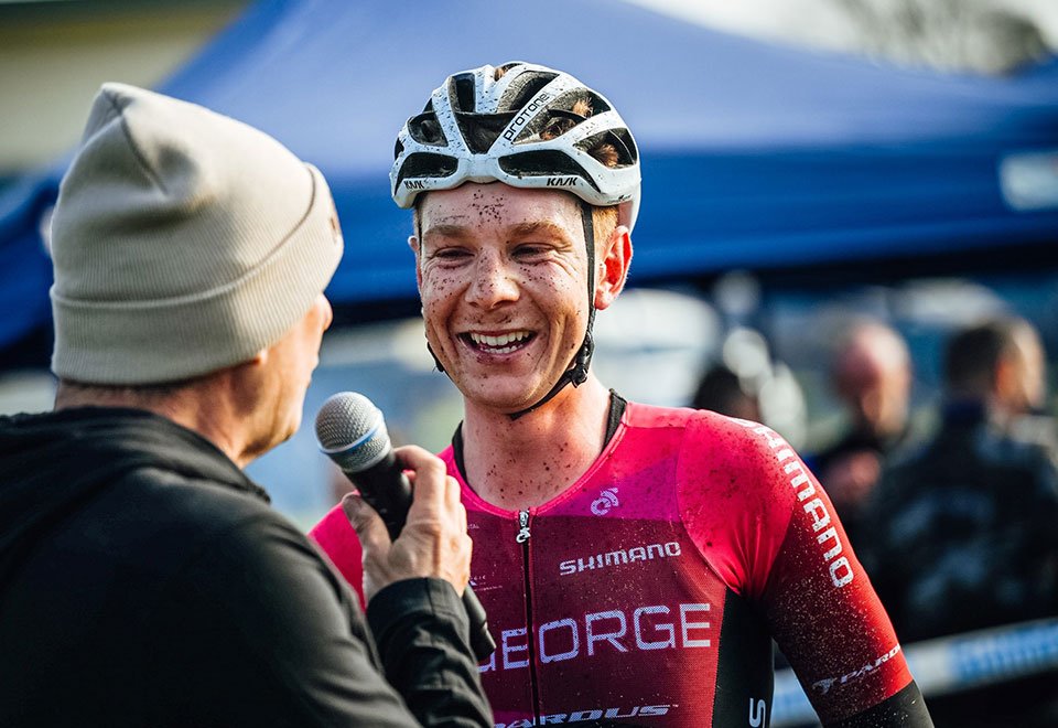Connor Sens interviewé après une course gravel, casque et maillot GEORGE recouverts de boue, performance technique et engagement total avec ruban Burgh