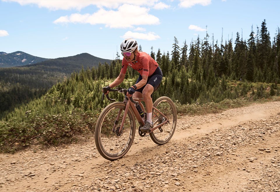 Justine Barrow en pleine course gravel sur piste caillouteuse, portant le casque blanc et le ruban de cintre Burgh, symbole de performance et d’endurance